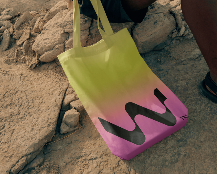 A person holding a tote bag with a gradient from yellow to pink, printed with a large black wavy “W” logo, resting on rocky terrain.